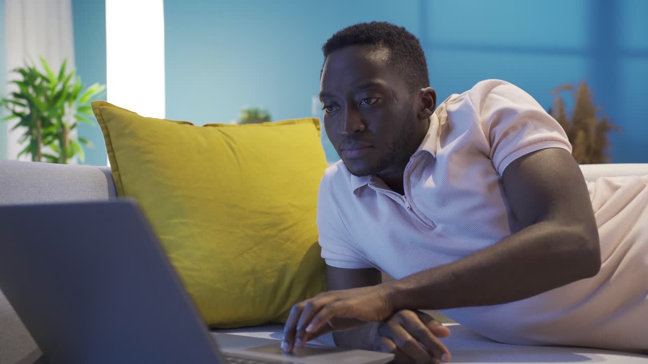 African young man using laptop lying on sofa at home at night.