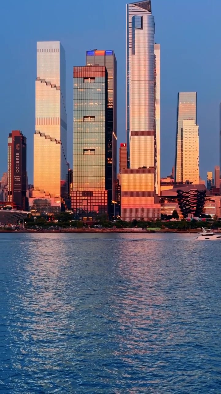 Modern skyscrapers of Hudson Yards reflecting sunset, New York City. Glass towers glow with orange light over blue water during dusk, vertical telephoto view.
