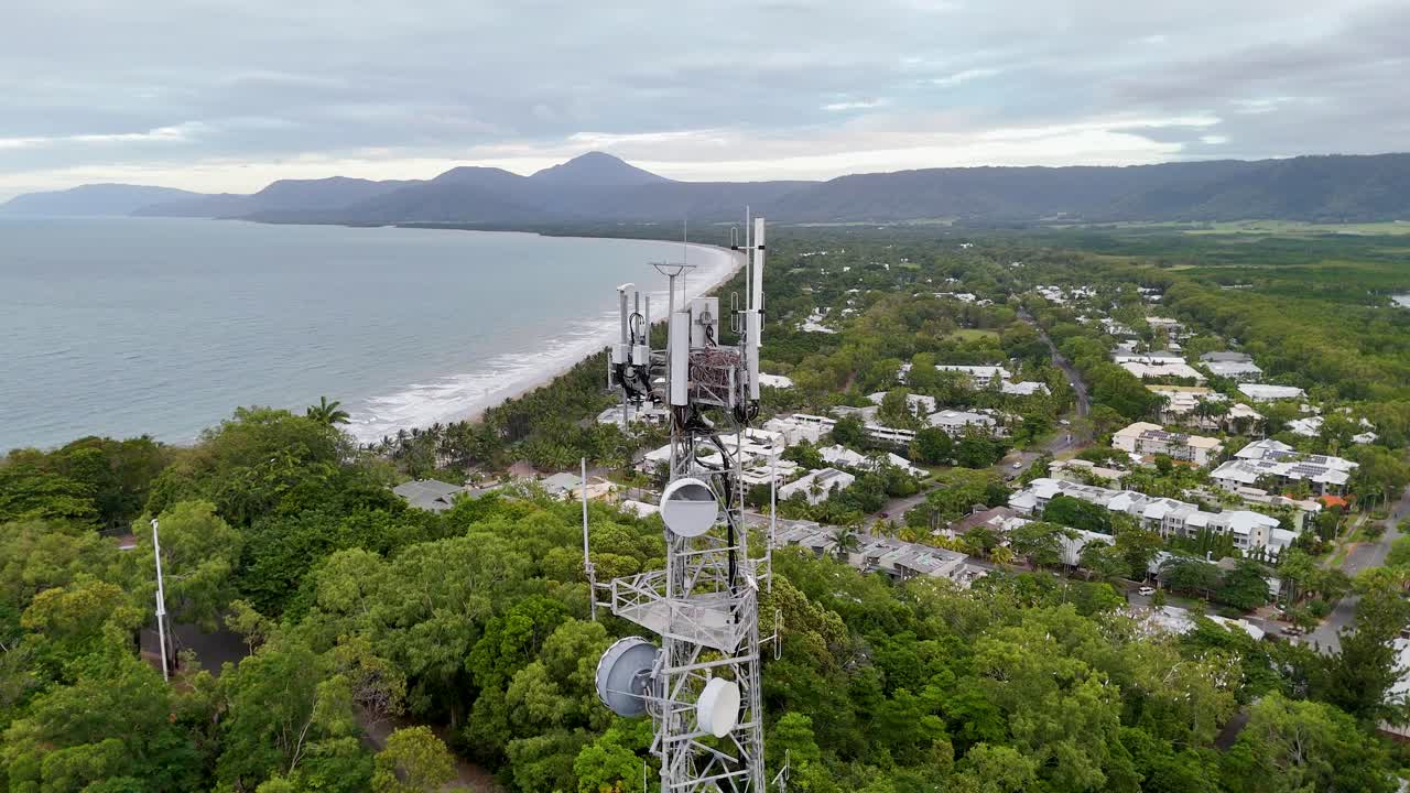 Drone footage captures a coastal town with lush greenery, a communication tower, and expansive ocean views under overcast skies