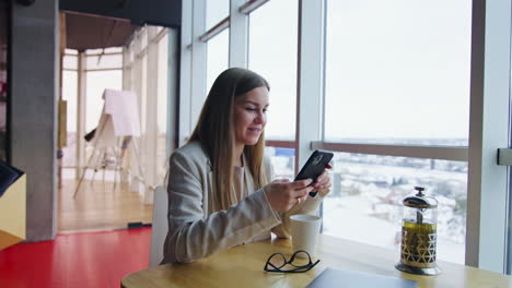 smiley lady sitting at table with cup of tea. cheerful woman looks at the phone and speaks. cityscape at backdrop.