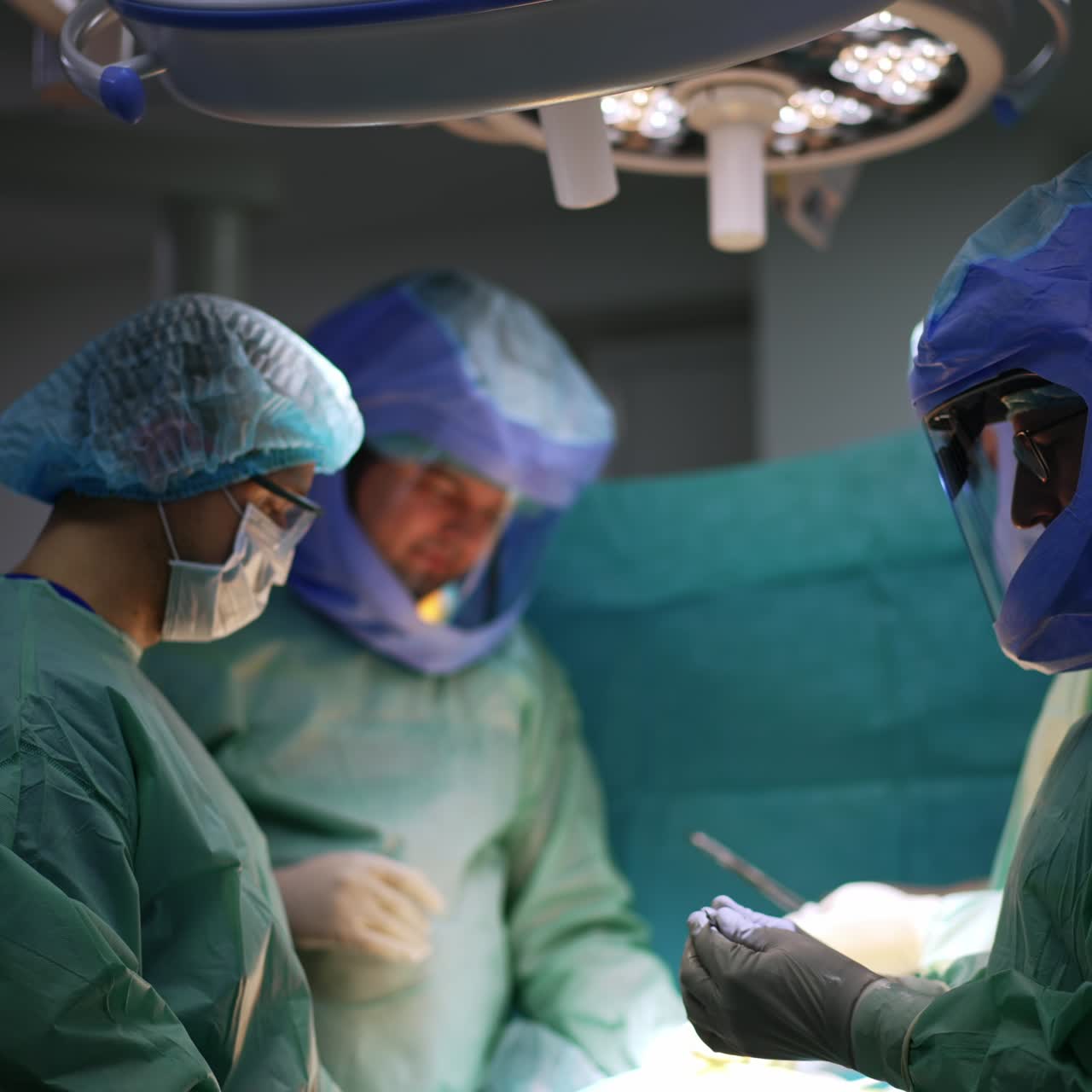 Surgical team in protective suits working under the bright lamp in surgery room. Medical professional cooperating at leg operation