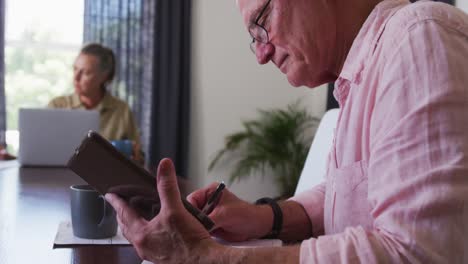 un anciano caucásico tomando notas mientras usa una tableta digital en casa