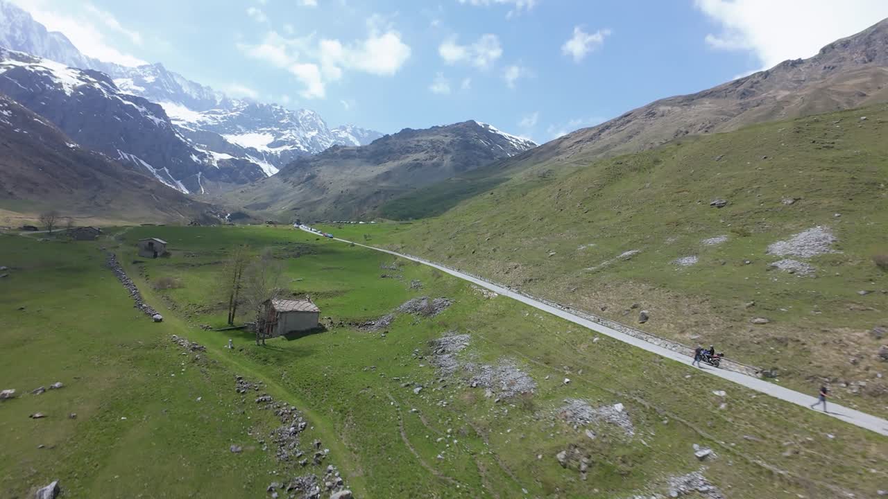 Filmato dinamico di un drone FPV che riprende un motociclista mentre percorre una lunga strada tortuosa in una valle ad alta quota circondata da cime innevate