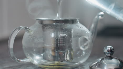 close up of person pouring hot water into glass kettle with filter as steam rises, showing herbal tea brewing with focus on transparent kettle and metal filter