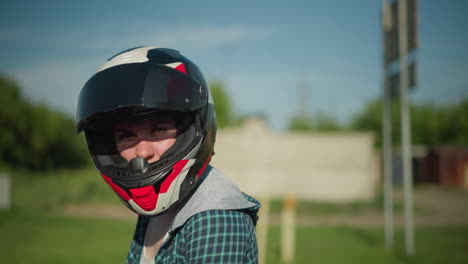 una motociclista femenina en equipo completo ajusta su casco levantando y bajando la visera, lleva un guante de protección negro, y un fondo borroso de árboles y un edificio es visible detrás de ella