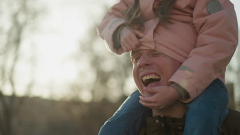 una niña con una gorra rosa y una chaqueta se sienta en el cuello de su padre, con su cabeza bien metida en su chaqueta. la niña juega tirando de la chaqueta de su cabeza mientras ambos sonríen brillantemente