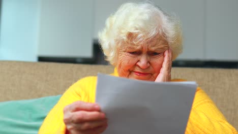 shoked amazed old woman white gray-haired sitting on the sofa in living room with bills