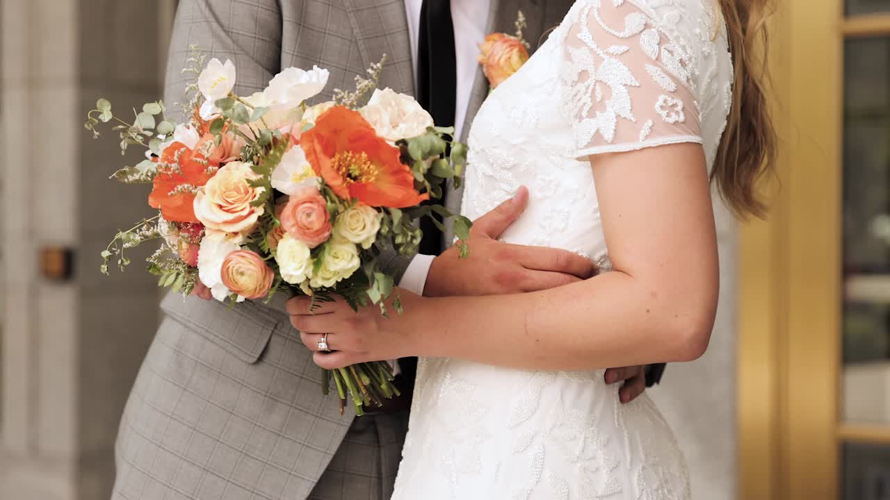 Primo piano di sposi che si abbracciano in abiti da sposa, con lei che tiene in mano un bouquet di fiori arancioni, rosa e bianchi, all'aperto 1080p 60fps