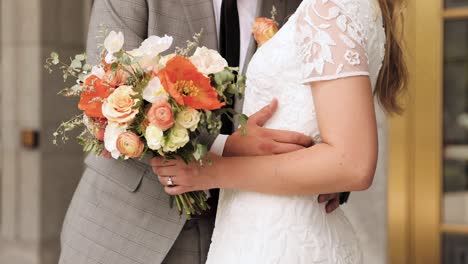 Close-Up-of-Bride-and-Groom-Hugging-in-Wedding-Attire-Holding-Orange,-Pink,-and-White-Flower-Bouquet-in-Her-Hands-Outdoors-1080p-60fps