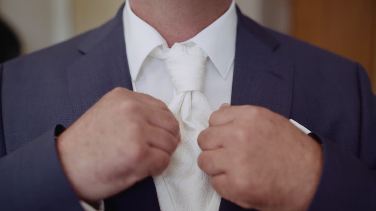 Close Up of groom adjusting patterned white tie on white shirt with blue suit