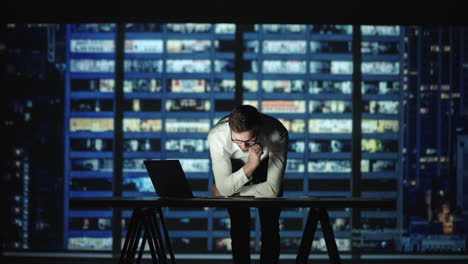 A young man stands at a table and looks at the graphs lying on the table and conducts a brainstorming session. Management of the company. Work at night. Creation and planning of the company&#039;s work