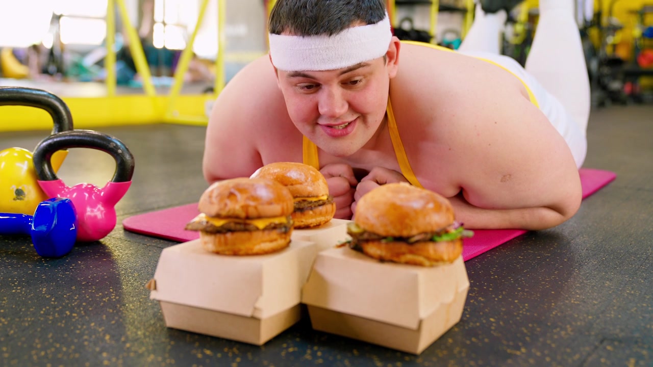 Uomo in sovrappeso su un tappetino da yoga tentato da cheeseburger in palestra