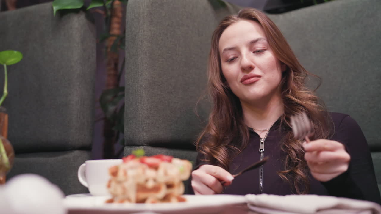 Close view of young woman in black top removing cutlery from folded napkin on wooden table, preparing to eat, in front are stacked waffles topped with strawberries and coffee in cozy cafe