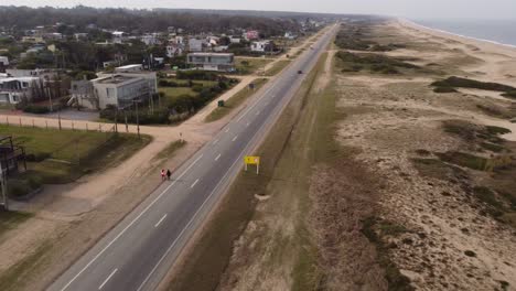 Toma-Aérea-De-Dos-Mujeres-Caminando-Al-Lado-De-La-Carretera-Costera-Cerca-De-La-Playa-En-Maldonado,-Uruguay