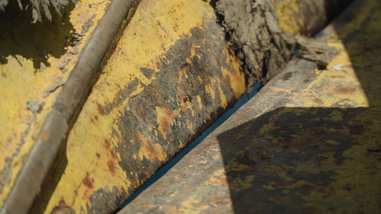 Rustent gult metalmaskineri tekstureret nærbillede, afskallende maling og mudder i sprækker, detalje af tungt udstyr med slid på bolt og flange, sollys kaster dybe skygger, barsk konstruktion.