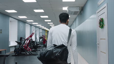 back view of young man walking out of gym hallway carrying duffel bag over shoulder after intense workout, gym equipment and other patrons visible in blurred background under bright ceiling lights