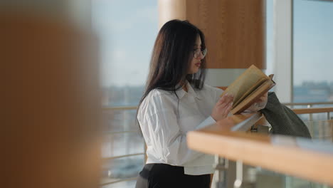 side view of young woman in white shirt reading book attentively while leaning on glass railing in sunlit modern interior, jacket draped beside her, cityscape visible through large windows