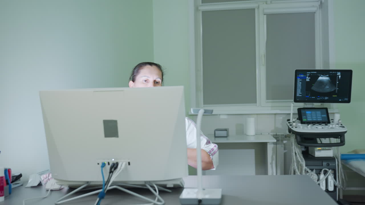 Female consultant in white uniform seated in front of desktop computer in modern clinical office focuses attentively on screen while scanning data with eyes near ultrasound equipment