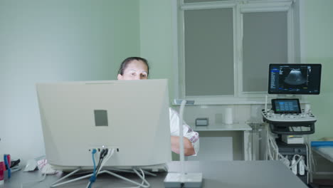 female consultant in white uniform seated in front of desktop computer in modern clinical office focuses attentively on screen while scanning data with eyes near ultrasound equipment