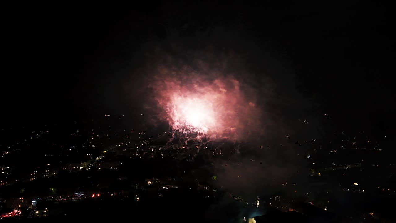 Close up aerial drone view of a bright pink and green firework explosion filling the frame with thick smoke and light over a dark city background during a festival
