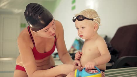 young mother and her cute little blonde child sitting by the swimming pool and preparing for the swimming lesson. happy mom