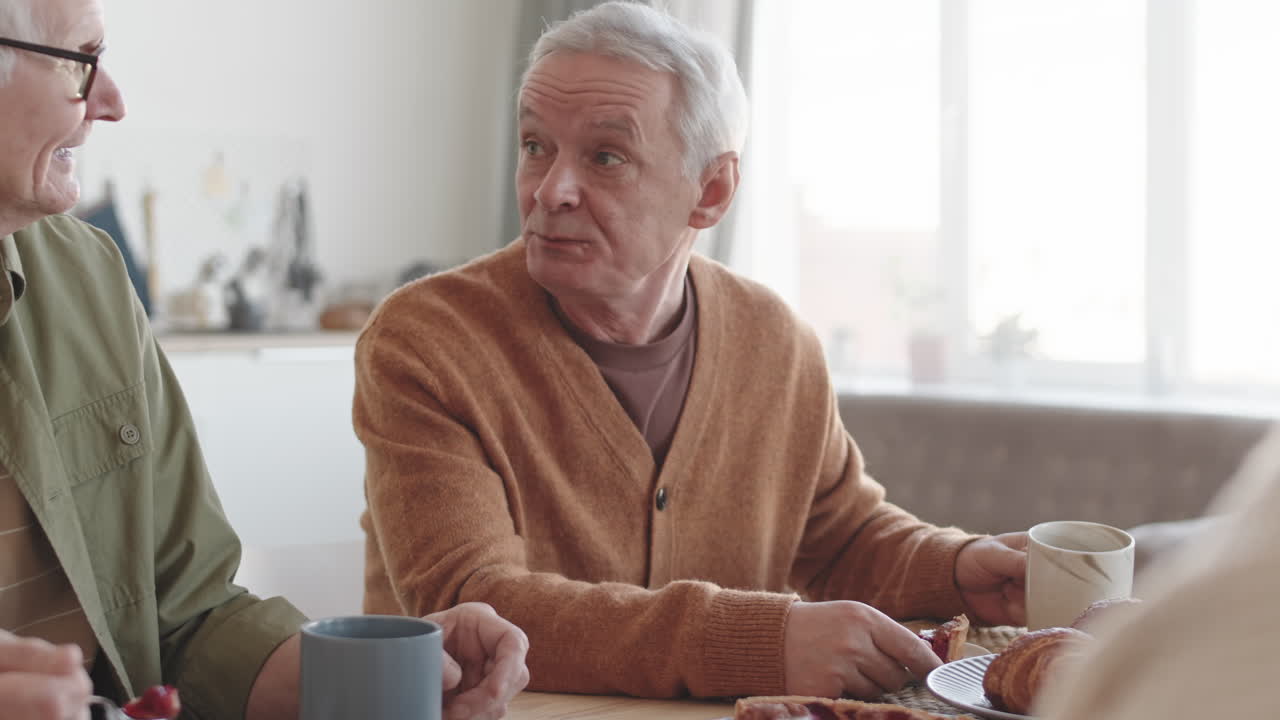 Tilting up of two senior Caucasian men sitting at table at home, eating sweet cake, drinking tea, talking to unrecognizable people