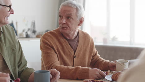 tilting up of two senior caucasian men sitting at table at home, eating sweet cake, drinking tea, talking to unrecognizable people