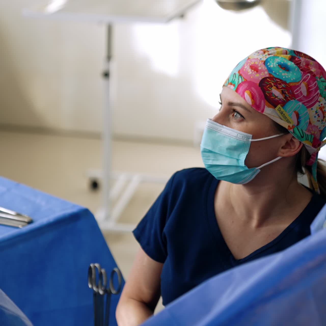 Female surgeon in colorful cap and mask performing operation. Doctor looks attentively aside on the screen showing procedure