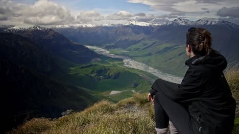 Girl-looking-over-a-beautiful-green-valley-with-mountains-with-a-river-running-through-on-a-bright-sunny-day-on-a-grassy-hill,-drifting-handheld-wide-medium-shot