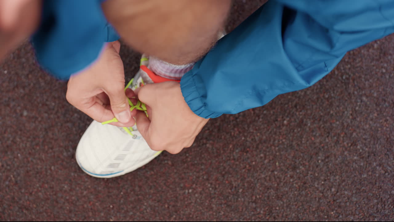 Corridore che si allaccia le scarpe da ginnastica in pista, primo piano dall'alto delle mani che stringono i lacci al neon, manica della giacca blu, superficie in gomma ruvida, rituale pre-corsa mirato e dettaglio di pioggia leggera, atleta che si prepara
