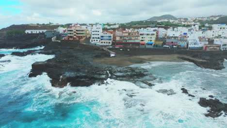 Vista-Aérea-En-órbita-Sobre-La-Playa-Y-Casas-En-La-Costa-De-El-Puertillo-En-El-Norte-De-La-Isla-De-Gran-Canaria-Y-Con-Grandes-Olas-Golpeando-La-Costa