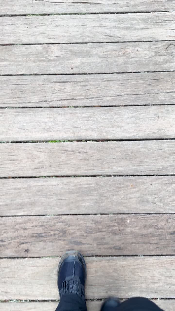 Vandrerens ben og støvler går stabilt på en solbeskinnet træpromenade, set fra et håndholdt nedadgående perspektiv.