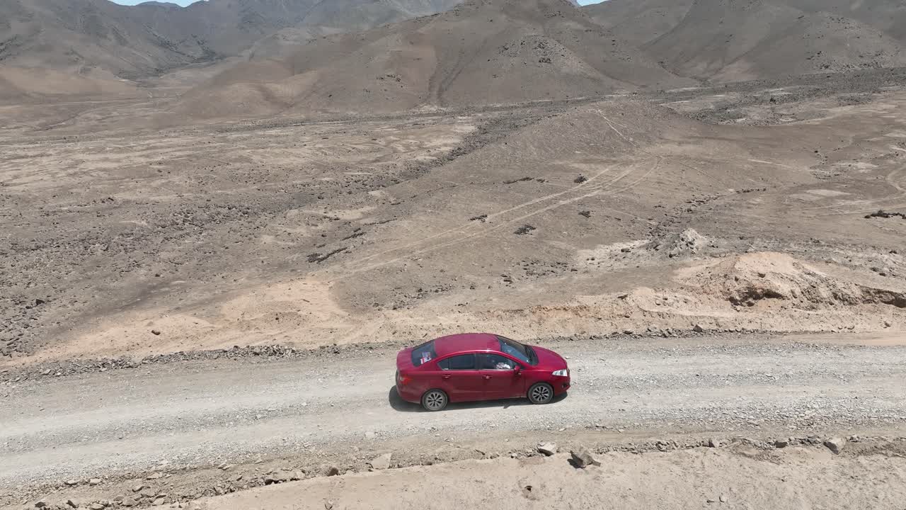 toma panorámica aérea de un vehículo rojo conduciendo en el desierto de perú hacia la ciudad santa de caral en una carretera polvorienta con colinas, piedras y montañas en el fondo en un día soleado