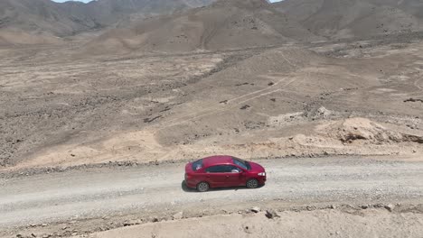 Luftschwenk-Eines-Roten-Fahrzeugs,-Das-An-Einem-Sonnigen-Tag-In-Der-Wüste-Von-Peru-In-Richtung-Der-Heiligen-Stadt-Caral-Auf-Einer-Staubigen-Autobahn-Mit-Hügeln,-Steinen-Und-Bergen-Im-Hintergrund-Fährt