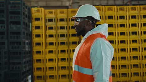 a man with black skin in safety glasses in a white helmet and a uniform with an orange vest walks along huge shelves with plastic boxes at a waste recycling and sorting plant