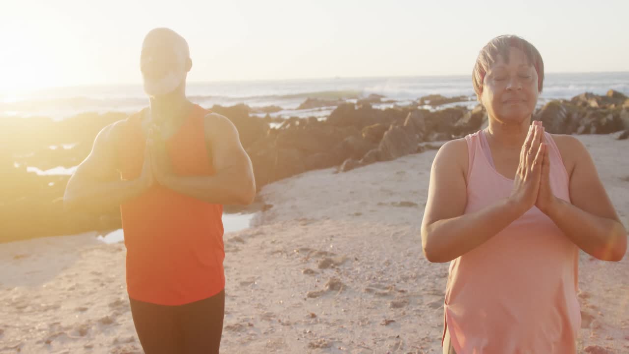 Coppia anziana afroamericana felice che fa yoga, meditando in spiaggia, al rallentatore