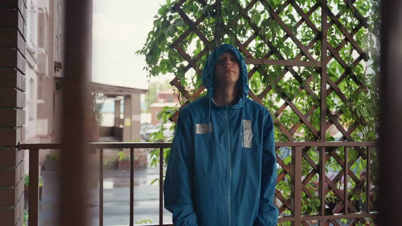 uomo con impermeabile blu sul balcone che guarda verso l’alto sotto una pioggia costante, cappuccio tirato su, gocce d’acqua sul tessuto, traliccio con edera dietro, strada bagnata e auto parcheggiate sotto, luci bokeh ambientali