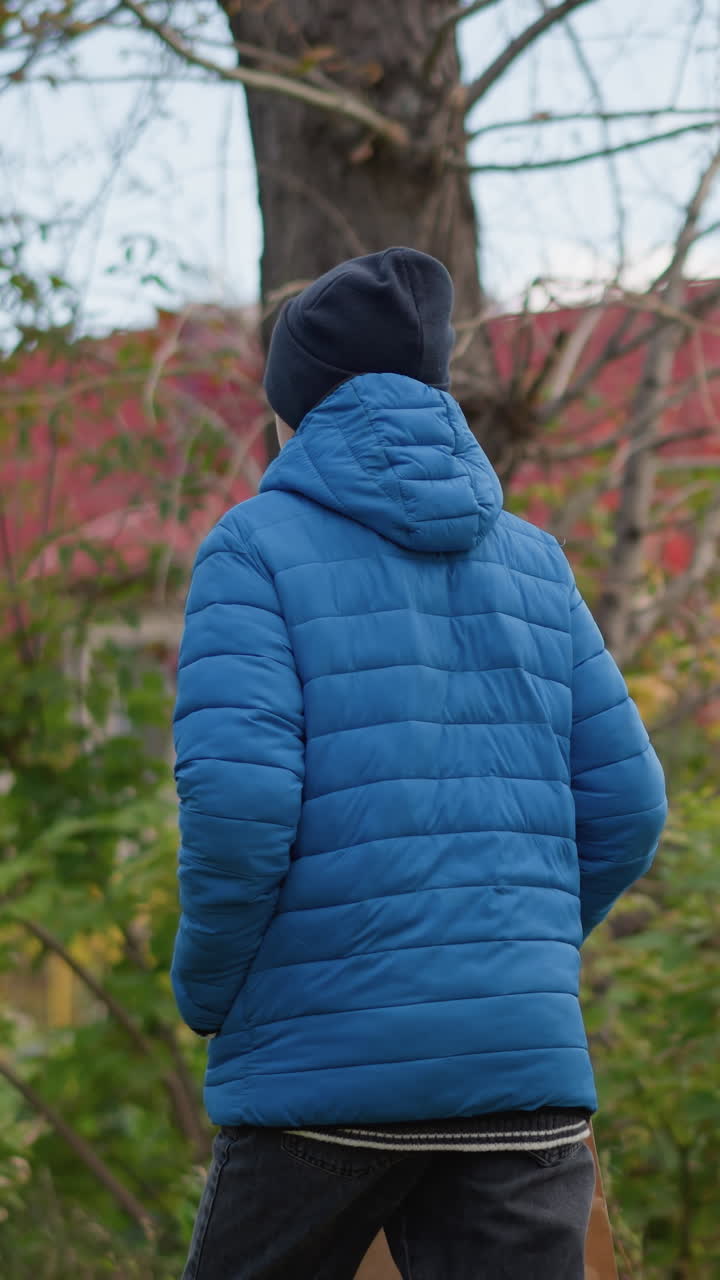 Back view of person wearing blue jacket and black beanie walking outdoors with paper bag, surrounded by lush greenery and colorful autumn foliage, with distant view of red-roofed building