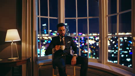 elegant man in a suit drinking coffee or tea by a window with a city night view