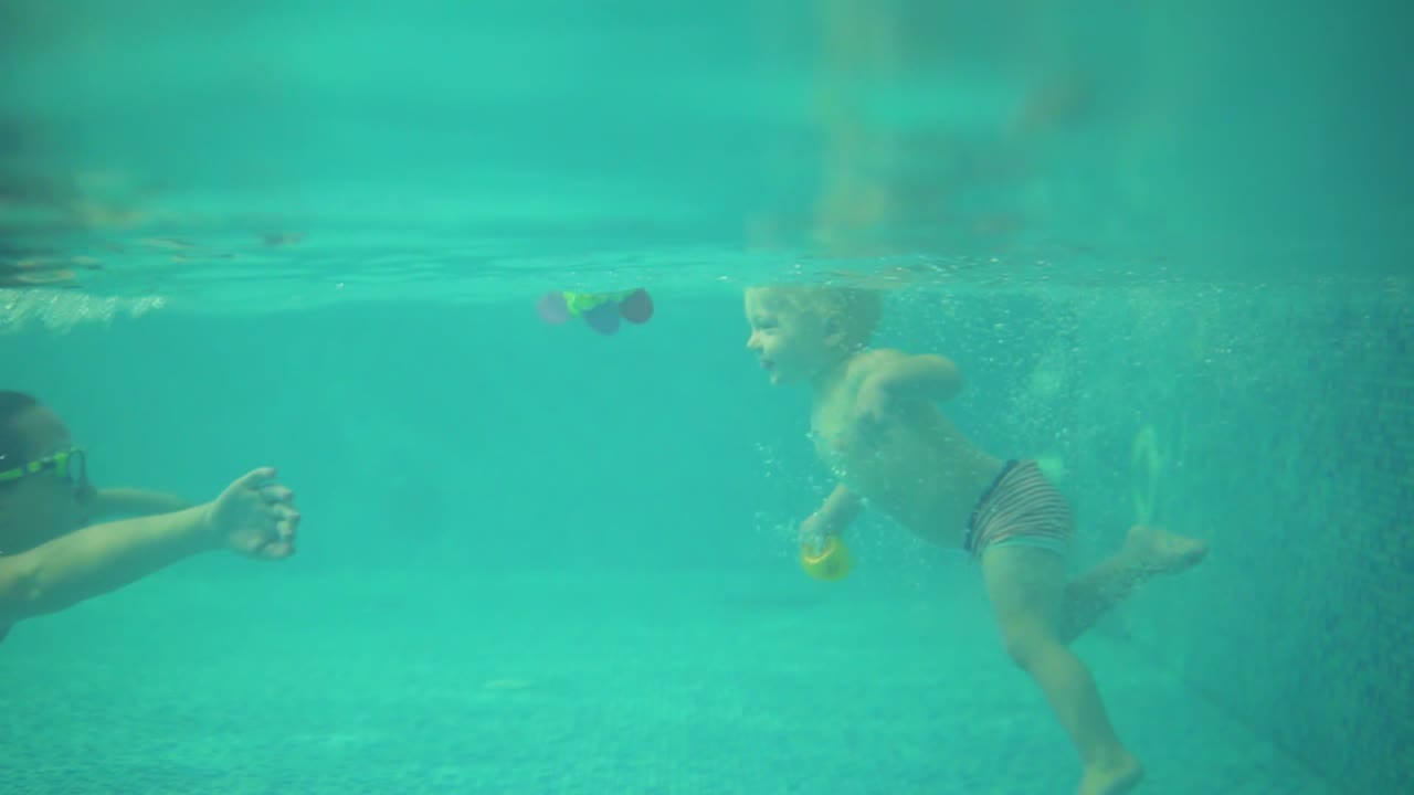 En undervandsoptagelse af en sød blond toddler, der svømmer under vandet sammen med sin mor i swimmingpoolen.