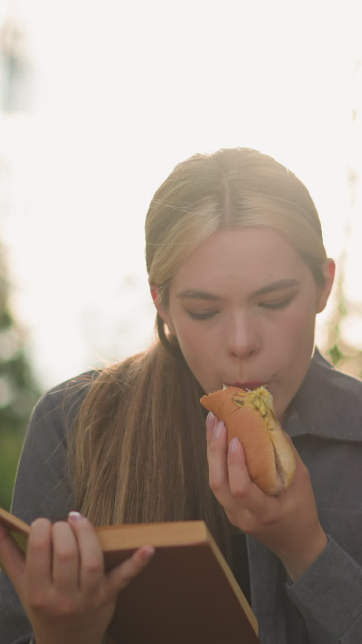 dama de camiseta gris disfrutando de un bocadillo mientras lee un libro al aire libre, sentada en un banco del parque, rodeada de árboles y vegetación con fondo borroso, la suave luz del sol ilumina