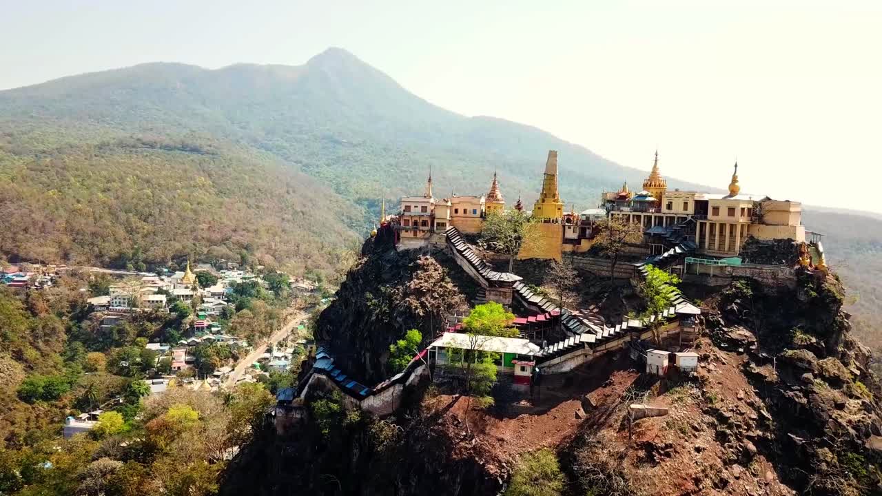 A stunning view of the Nat Temple in Myanmar, nestled atop a rugged mountain ridge with golden spires and panoramic scenery, surrounded by lush forests and nearby village dwellings