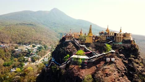 una impresionante vista del templo de nat en myanmar, ubicado en lo alto de una escarpada cresta montañosa con torres doradas y paisajes panorámicos, rodeado de frondosos bosques y viviendas de aldeas cercanas