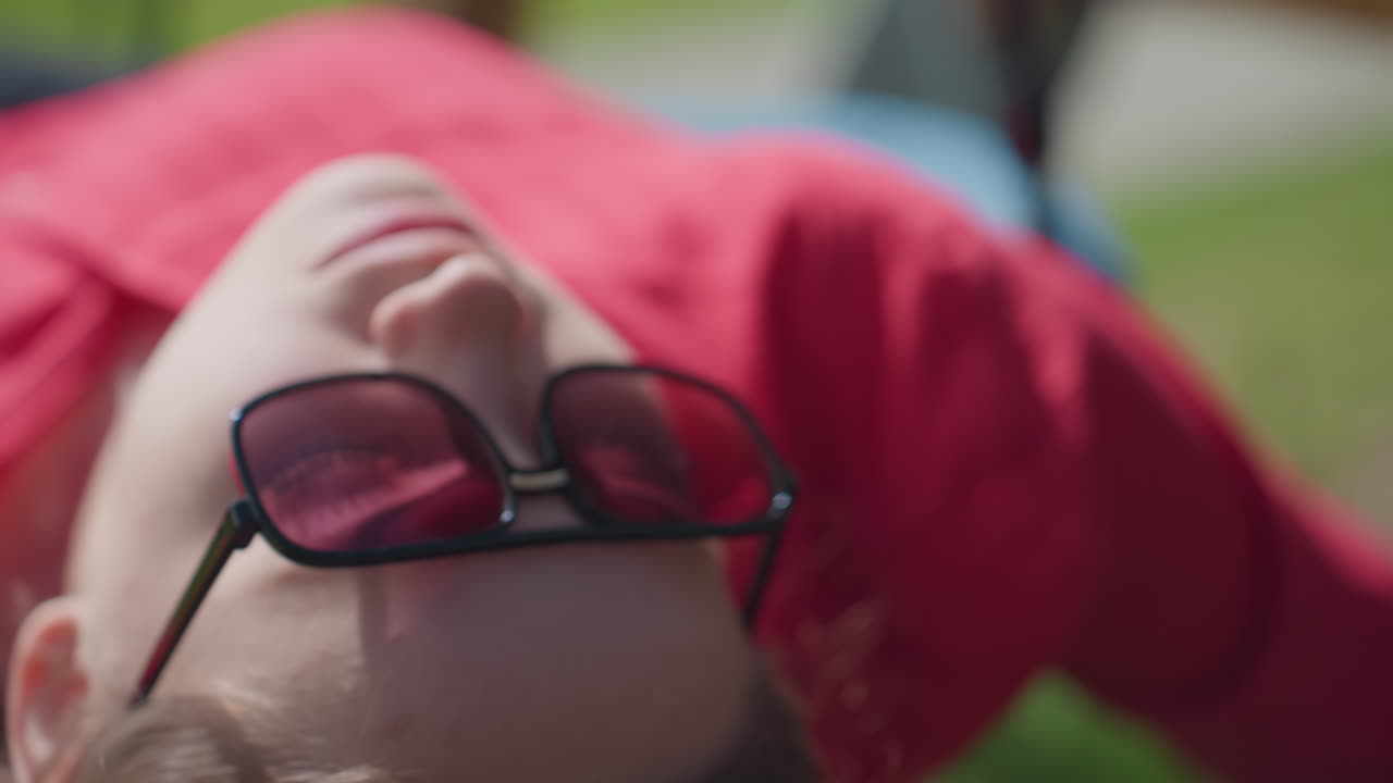 Close up of woman wearing sunglasses while gently swinging under sunlight, her head tilts back in relaxed position, eyes closed behind tinted lenses, surrounded by outdoor greenery and warm