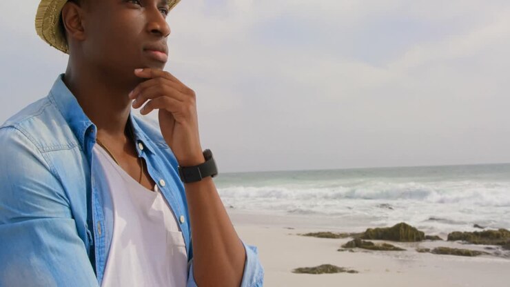 Vista laterale di un uomo afroamericano in piedi con la mano sul mento in spiaggia 4k