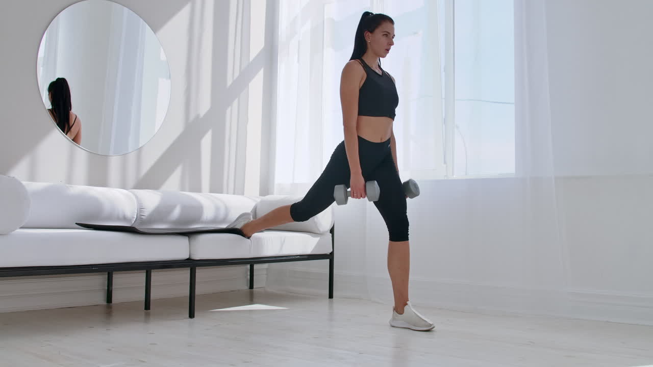 Una bruna in abbigliamento sportivo nero in un appartamento bianco esegue uno split squat con manubri tra le mani, appoggiando il piede sul divano.