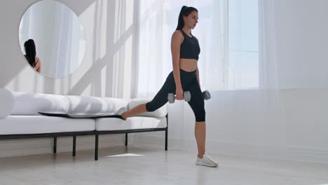 brunette in black sportswear in a white apartment makes a split squat with dumbbells in her hands leaning on the sofa with her foot.