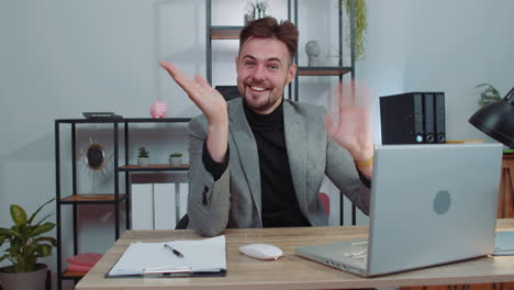 hombre de negocios trabajando en una computadora portátil sonriendo amigablemente a la cámara y agitando las manos saludando en la oficina