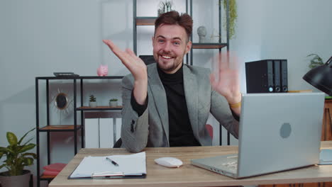 businessman working on laptop smiling friendly at camera and waving hands gesturing hello at office