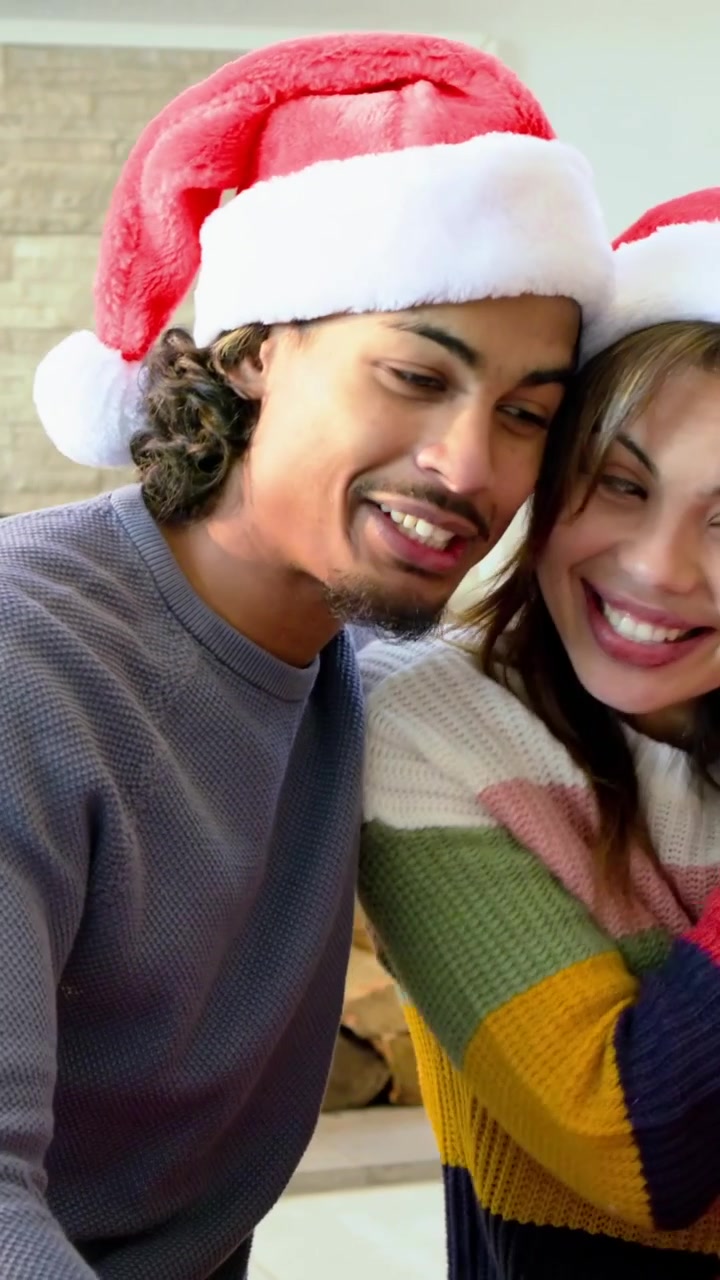 Video vertical: Hombre con gorro de Papá Noel extendiendo el brazo hacia una mujer con suéter para una selfie en casa con un teléfono.